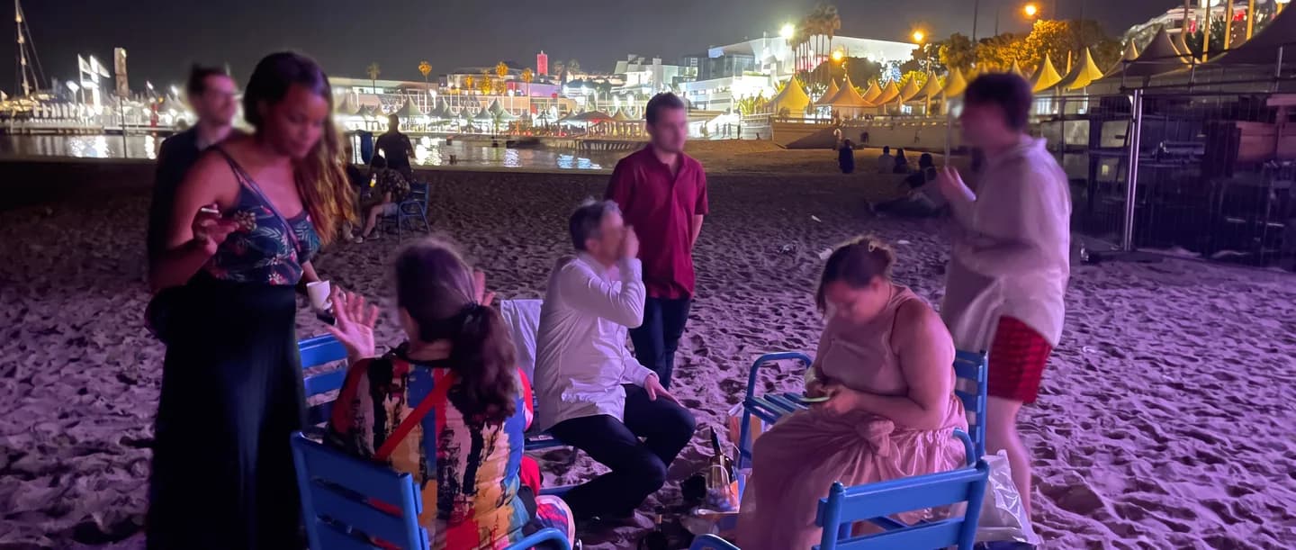 Un groupe d'amis assis sur des chaises de plage, le soir, devant les lumières de la Croisette à Cannes.